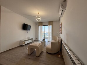 A modern living room with a flat-screen TV, a white console table, a beige sofa, a round coffee table, and a floor lamp. - Malaga 2 Bright Luxury 1BR Central Location (La Marsa)