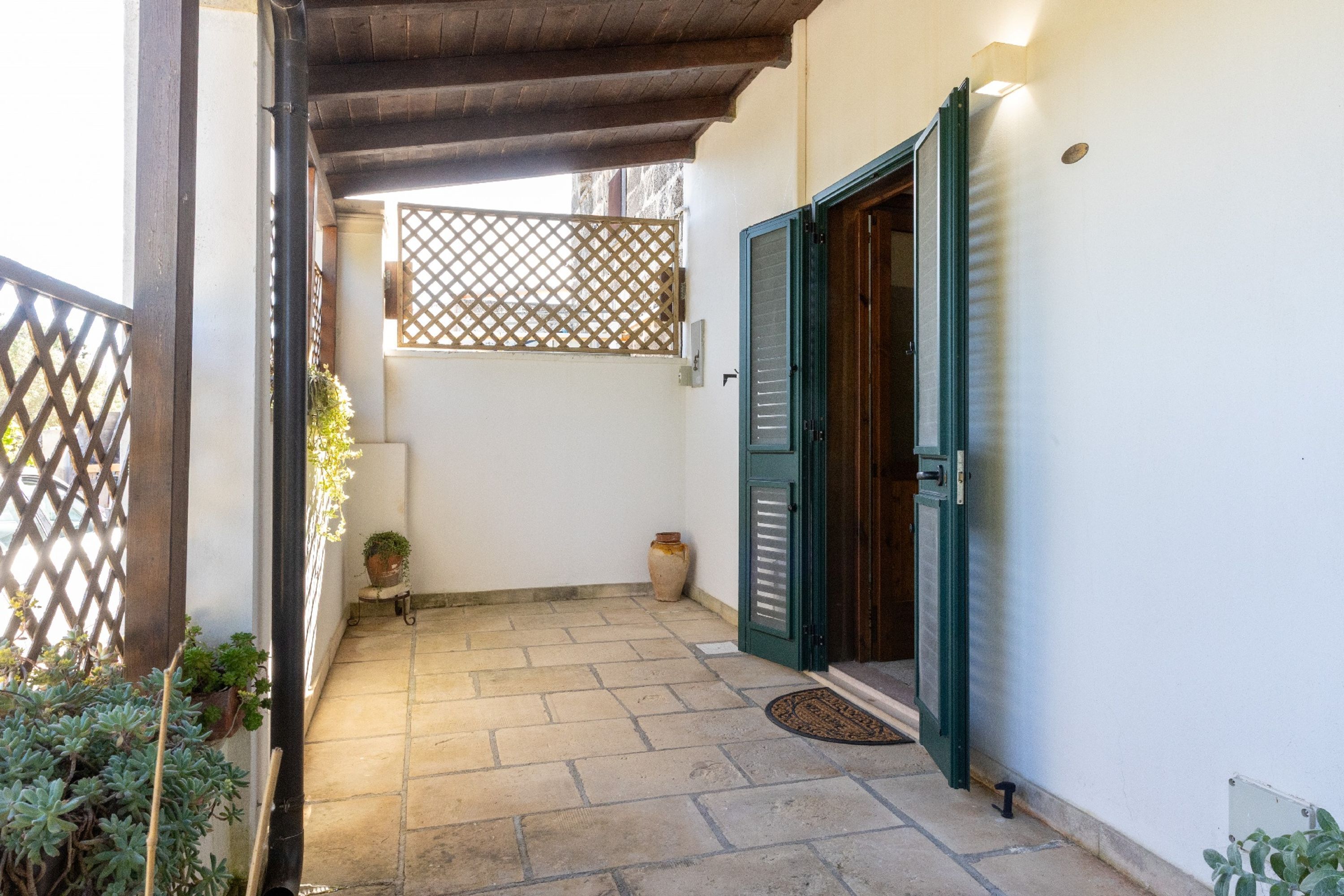 Un pequeño patio con piso de baldosas, una pérgola de madera, una puerta verde y plantas en macetas.