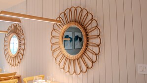 Ett rum med en väggmonterad spegel med ett solliknande motiv, ett träbord med stolar och en inramad tavla på väggen.