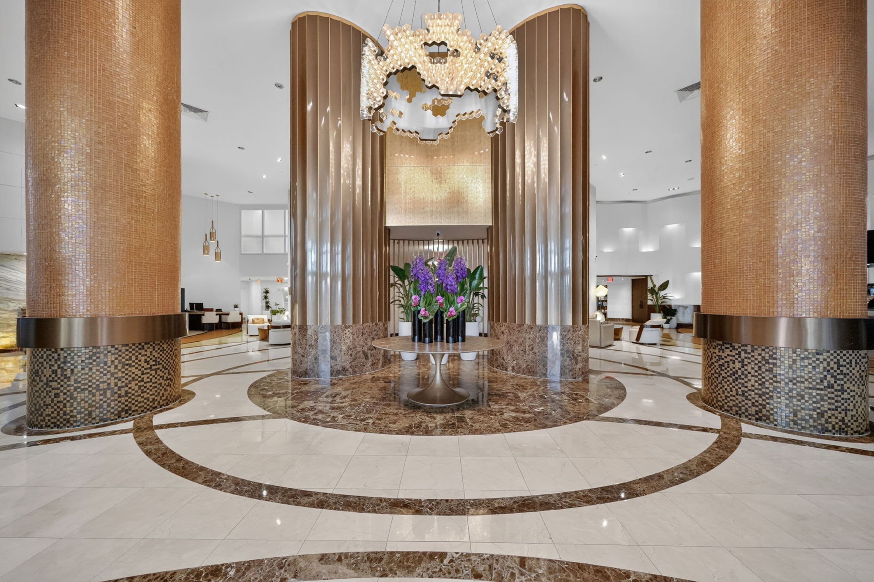 A spacious lobby with a central reception desk, a decorative chandelier, and a floral arrangement.