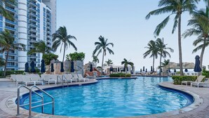 A swimming pool with lounge chairs and umbrellas, surrounded by palm trees and a hotel building. - Luxury Beachfront Escape Sunny Isles (Sunny Isles Beach)
