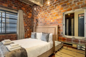 Ein Hotelzimmer mit Backsteinwand, einem breiten Bett mit vielen Kissen, einem Spiegel und einem Fenster mit Vorhängen.