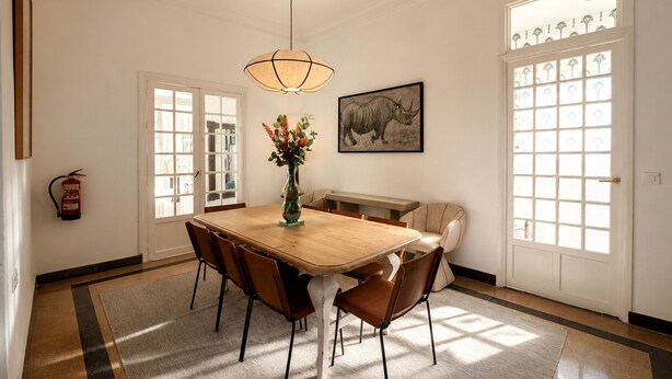 Une salle à manger avec une table en bois, six chaises, un vase de fleurs, un tableau au mur et un extincteur.