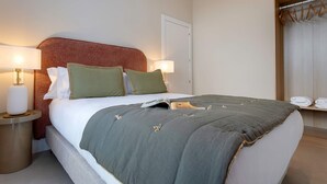 Una cama bien tendida con una manta gris y dos almohadas, una mesita de noche con una lámpara y una ventana con cortinas.