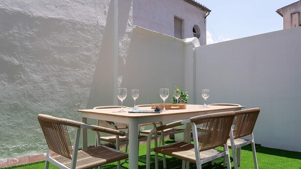 Una mesa con cuatro sillas, una bandeja y cuatro copas de vino, frente a una pared blanca con vista a una masa de agua.