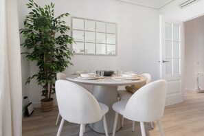 Et spiseområde med et rundt bord, hvide stole, en potteplante og et vindue med udsigt.