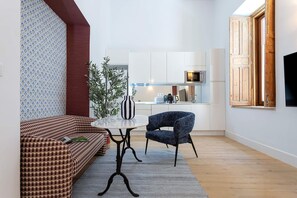 Ein modernes Wohnzimmer mit einem karierten Sofa, einem runden Tisch und einem blauen Sessel. Es gibt eine Küchenzeile mit weißen Schränken und einer Mikrowelle.