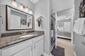 A bathroom with a granite countertop sink, a large mirror with lights, a glass shower enclosure, and a bed visible through an open door. - #217 Pointe - 2 Bedroom Lakefront Condo at Mariners SML (Bedford County)
