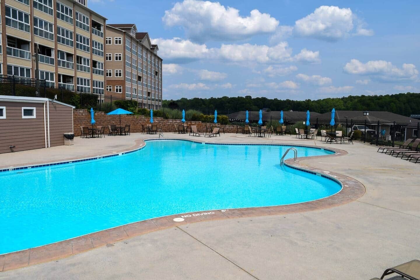 Une piscine entourée de chaises longues et de parasols, elle-même bordée d’un immeuble à plusieurs étages et doté de nombreuses fenêtres.
