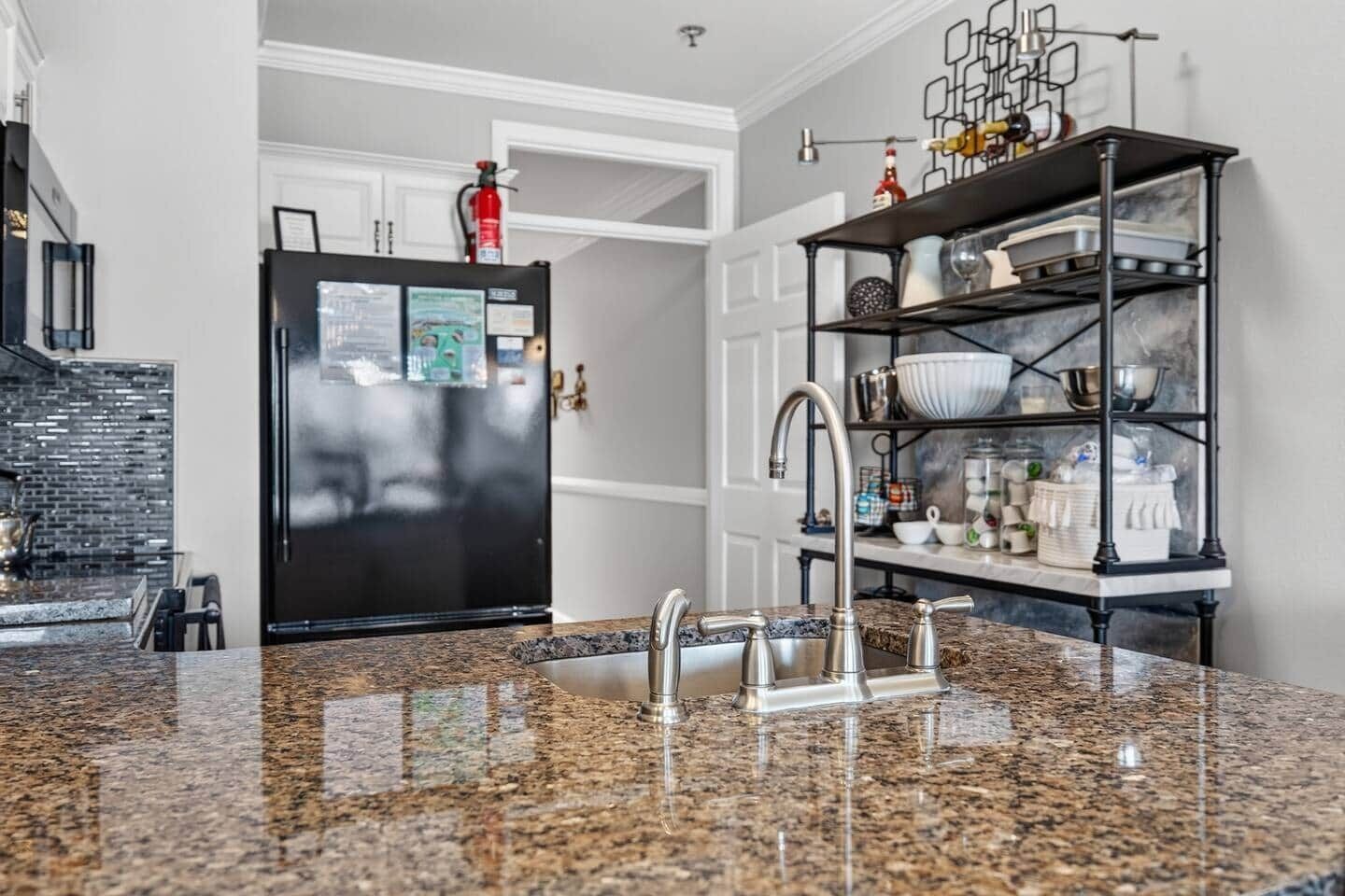 Eine Küche mit Granit Arbeitsplatten, einer Edelstahl-Spüle, einem schwarzen Kühlschrank und einem schwarzen Regaleinheit mit Geschirr.