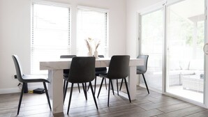 Comedor con una mesa blanca y cuatro sillas, ventanas grandes con persianas y vista al exterior.
