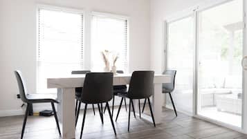Un coin repas avec une table blanche et quatre chaises, de grandes fenêtres équipées de stores et une vue sur l’extérieur.