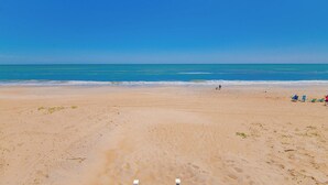 Ein Strand mit blauem Himmel, sandigem Ufer und einigen Personen sowie Strandliegen.