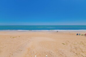 Ein Strand mit blauem Himmel, sandigem Ufer und einigen Personen sowie Strandliegen.