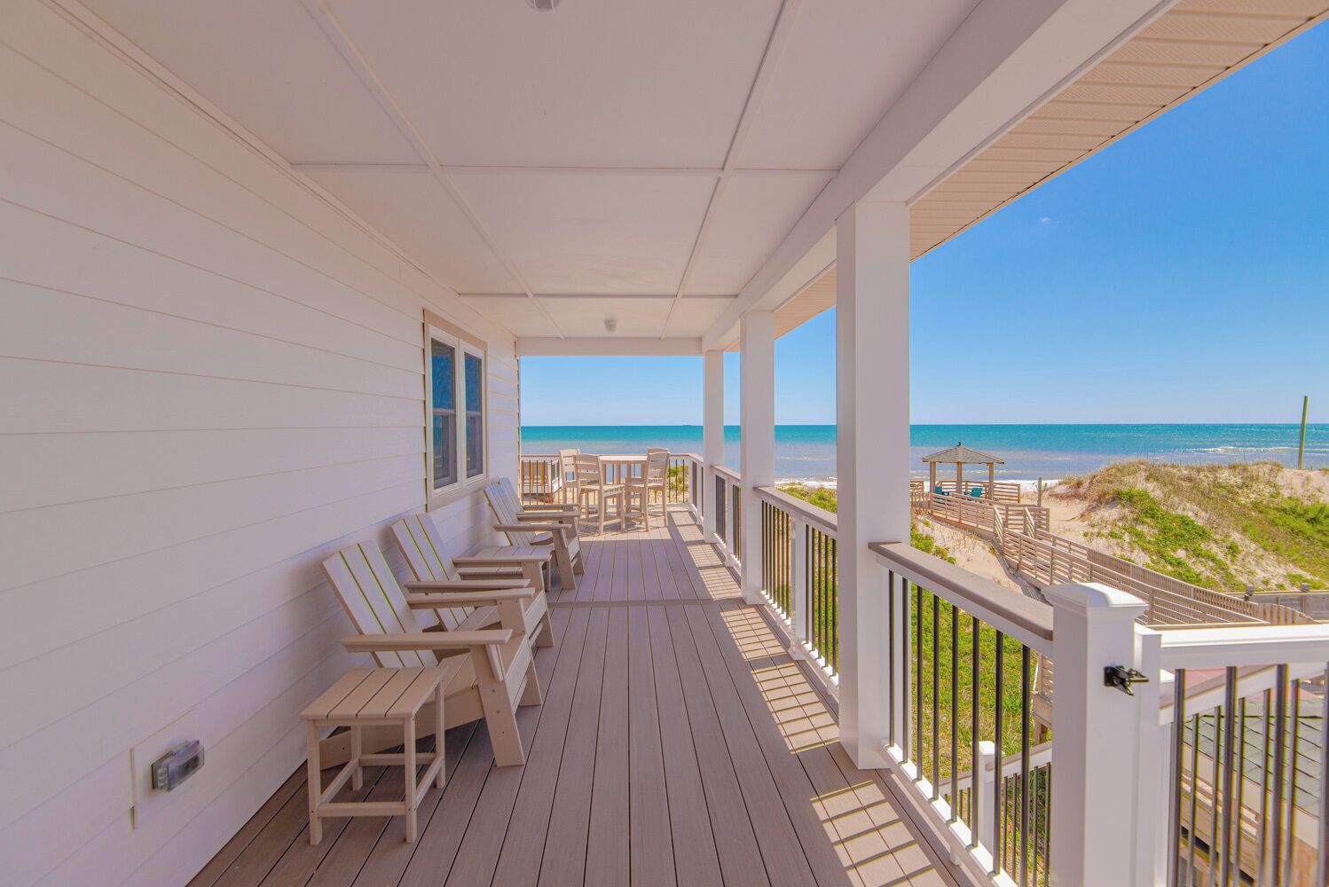 A covered porch with wooden chairs and tables, overlooking a beach with dunes and a clear sky.