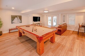 Ein Billardtisch in einem Raum mit Holzboden, einer Couch, einem Fernseher und einem Fenster mit Jalousien.