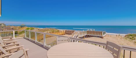 A beachside deck with a round table and chairs, wooden railings, and a view of the ocean.