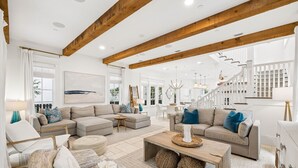 A spacious living room with a high ceiling and exposed wooden beams, a staircase leading to an upper level, and a large window providing natural light. - Walk to Beach and Square | Private Pool (Inlet Beach)