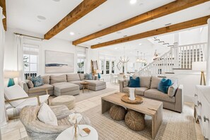Ein geräumiges Wohnzimmer mit hoher Decke und freiliegenden Holzbalken, eine Treppe, die zu einer oberen Ebene führt, und ein großes Fenster, das für natürliches Licht sorgt.