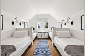 Ein Zimmer mit zwei Einzelbetten, einem kleinen Tisch mit Stauraum, einem Fenster und einem Deckenventilator.