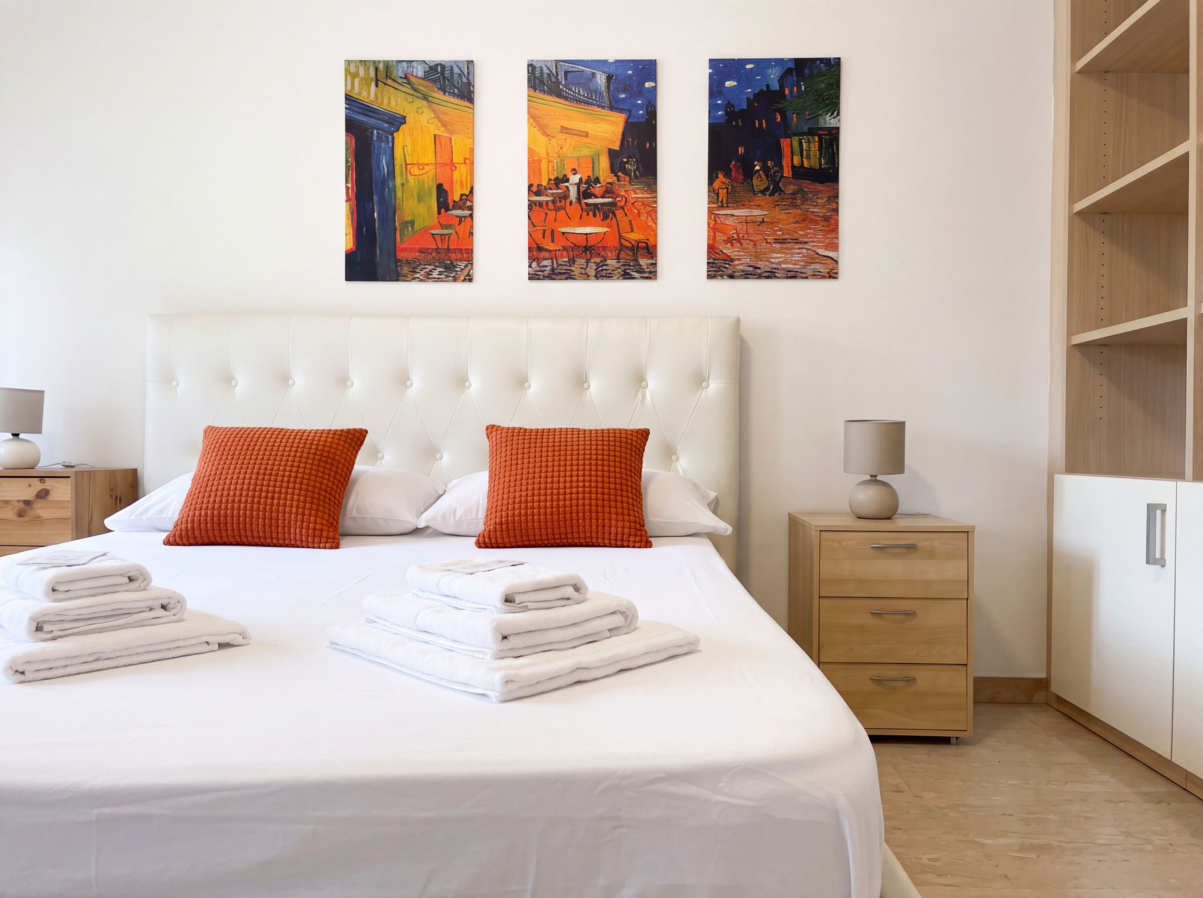 A hotel room with a large bed, two red pillows, a white headboard, a nightstand with a lamp, and three paintings on the wall.