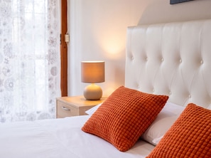 Ein ordentlich bezogenes Bett mit weißer Bettwäsche und zwei orangefarbenen Kissen, ein Nachttisch mit Lampe und ein Fenster mit durchsichtigen Vorhängen.