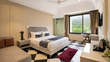 Habitación de hotel con cama, escritorio, silla, televisor y ventana con vista a vegetación.