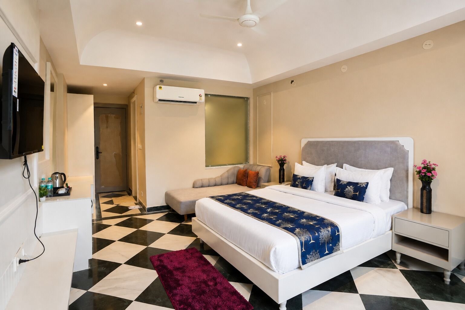 A hotel room with a checkered floor, a bed with white and blue bedding, a grey sofa, a TV mounted on the wall, and a ceiling fan.