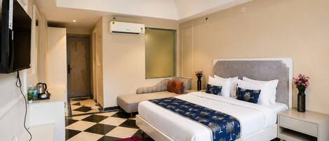 A hotel room with a checkered floor, a bed with white and blue bedding, a grey sofa, a TV mounted on the wall, and a ceiling fan.