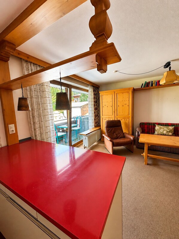 A kitchen with a red countertop, wooden ceiling beams, a brown leather chair, a wooden table, and a window with curtains.