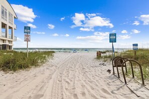 Ein sandiger Küstenpfad führt zum Meer, mit Menschen und Liegestühlen am Strand.