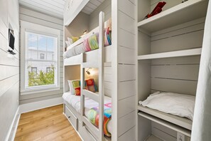 Ein Zimmer mit Fenster, einem Bett mit bunter Bettwäsche, einem Regal mit diversen Gegenständen und einer weißen Wand.