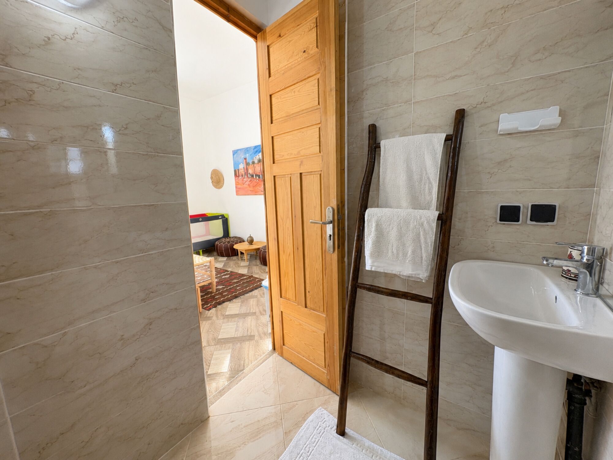 A bathroom with a wooden door, a wooden ladder, a white sink, and a tiled floor.