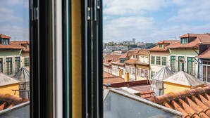 Vista de edifícios com telhados em terracota e molduras de janelas verdes, céu claro e uma paisagem urbana ao longe.