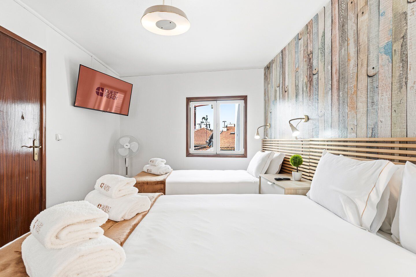 Habitación de hotel con cama, mesita de noche, ventilador, televisor montado en la pared y ventana con vista a edificios.