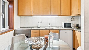Cozinha compacta com armários de madeira, micro-ondas, pia e área de jantar com mesa de vidro.