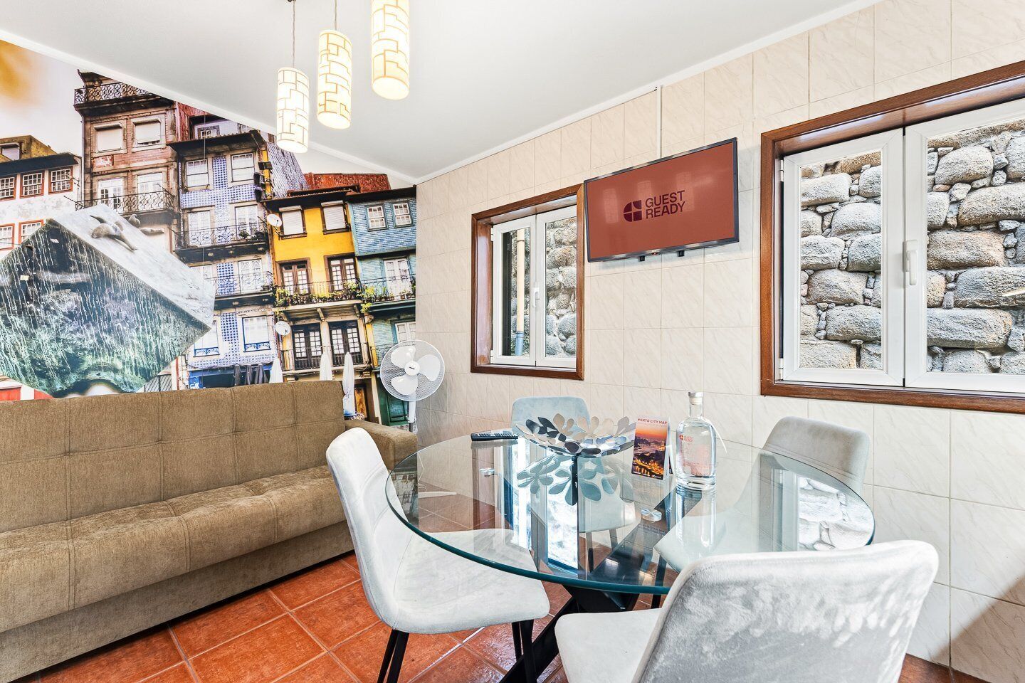 A hotel room with a sofa, a glass table, chairs, a fan, a TV, and a wall with a cityscape mural.