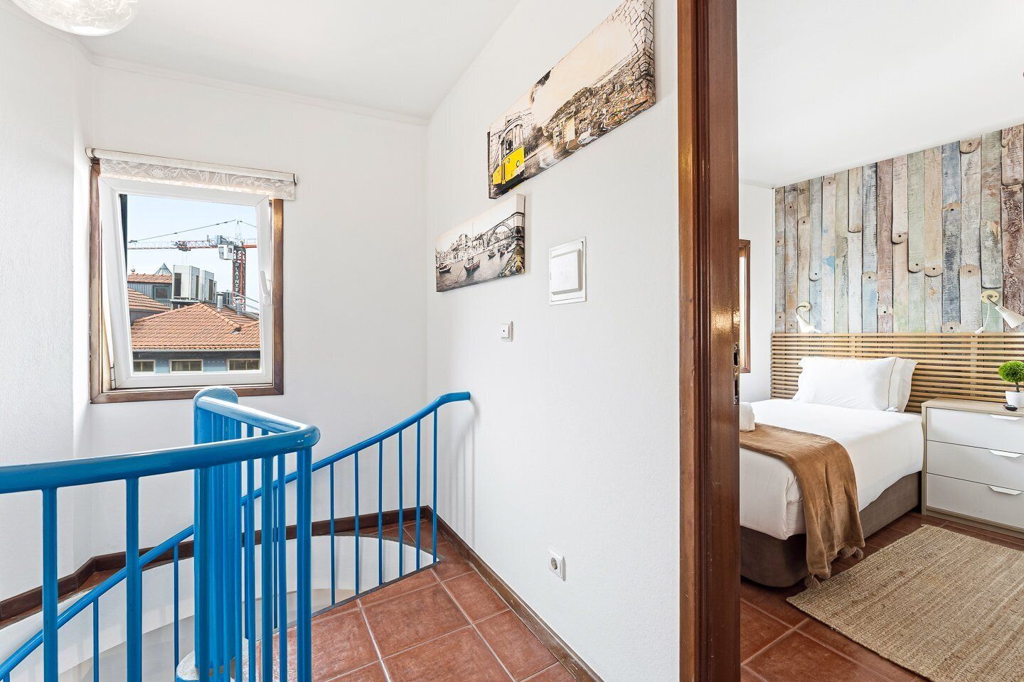 A hotel room with a bed, a nightstand, a window with a view of buildings, a staircase with blue railings, and a wall with a wooden plank design.