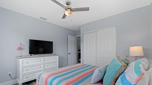 A bedroom with a bed, a TV, a dresser, a ceiling fan, and a door. - Dune Your Thing A&B (Fort Myers Beach)