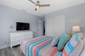 A bedroom with a bed, a TV, a dresser, a ceiling fan, and a door. - Dune Your Thing A&B (Fort Myers Beach)