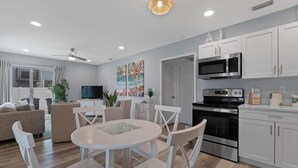 A modern kitchen with white cabinets, a microwave, and a stove. There is a dining area with a white table and chairs. - Dune Your Thing A&B (Fort Myers Beach)