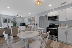 A modern kitchen with white cabinets, a microwave, and a stove. There is a dining area with a white table and chairs. - Dune Your Thing A&B (Fort Myers Beach)