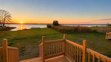 Una terraza de madera con barandilla que da a un césped y una vista del atardecer sobre el agua.