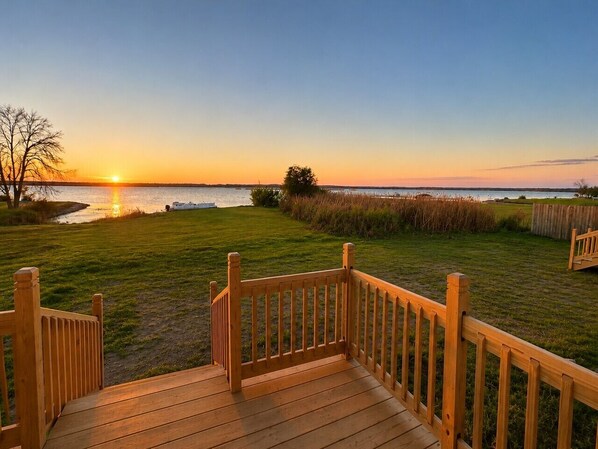 Una terraza de madera con barandilla que da a un césped y una vista del atardecer sobre el agua.