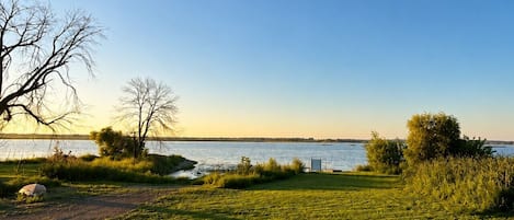 A grassy area with a view of a body of water, a wooden deck, and a fire pit.