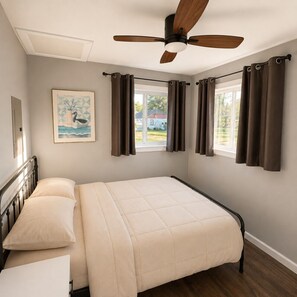 Un dormitorio con cama, ventilador de techo, ventana con cortinas y un cuadro en la pared.