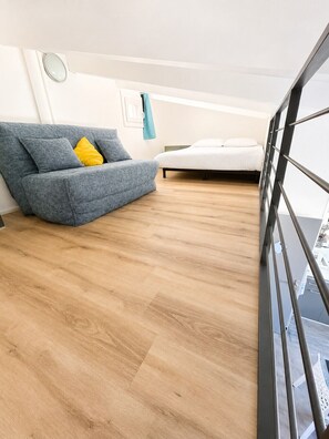 A modern living room with a sofa and a bed, wooden flooring, and a view through a window. - EUREKA 238  - Appartement -Piscine Terrasse - Plages à pied (Agde)