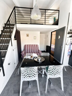 A compact living space with a dining table, chairs, a sofa, a staircase, and a hanging light fixture. - EUREKA 238  - Appartement -Piscine Terrasse - Plages à pied (Agde)