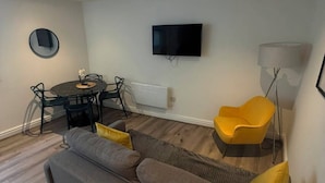 Ein modernes Wohnzimmer mit einer Couch, einem Esstisch mit Stühlen, einem flachbildfernseher an der Wand und einer Stehlampe.