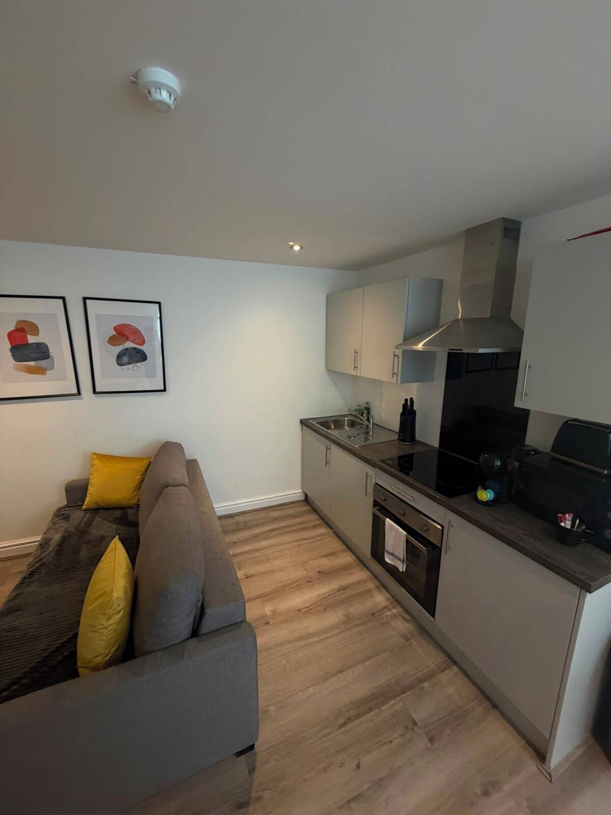 Een moderne keuken met een ingebouwde oven, een kookplaat en een afzuigkap. In de woonkamer staat een grijze bank met gele kussens.